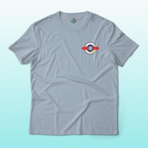 Puede incluir: Camiseta azul claro con un peque&ntilde;o gr&aacute;fico en el pecho. El gr&aacute;fico presenta un dise&ntilde;o rojo, blanco y azul con un acento amarillo. La camiseta tiene mangas cortas y cuello redondo.