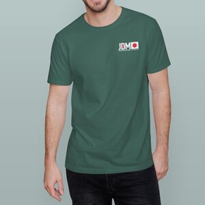 May include: A dark green t-shirt with the text "JDM" and a Japanese flag design on the upper left chest. The shirt is a crew neck style and is made of a soft material.