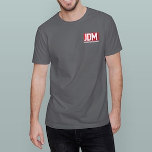 May include: A gray t-shirt with the text "JDM JAPANESE DOMESTIC MARKET" printed in red and white on the upper left chest. The shirt is short-sleeved and made of a soft material.