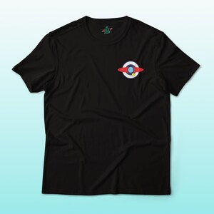 Puede incluir: Camiseta negra con un peque&ntilde;o gr&aacute;fico colorido de un planeta o nave espacial en el pecho. El gr&aacute;fico incluye elementos rojos, blancos, azules y amarillos. La camiseta tiene mangas cortas y cuello redondo.