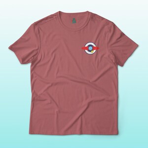 Puede incluir: Una camiseta de color rosa polvoriento con un peque&ntilde;o gr&aacute;fico colorido en el pecho izquierdo. El gr&aacute;fico presenta un c&iacute;rculo blanco con un c&iacute;rculo azul en el interior, una banda roja y un punto amarillo. La camiseta tiene mangas cortas y cuello redondo.