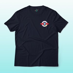 Puede incluir: Camiseta azul marino con un peque&ntilde;o gr&aacute;fico colorido en el pecho. El gr&aacute;fico presenta un anillo rojo y blanco con un centro azul y un punto amarillo. La camiseta tiene mangas cortas y cuello redondo.