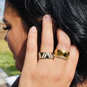 May include: Two gold rings on a finger. The top ring has a black and white geometric design. The bottom ring has a row of clear stones.