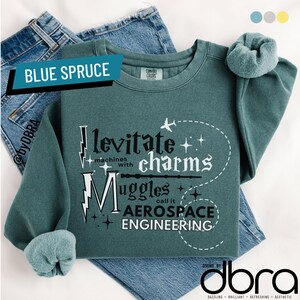 May include: Blue spruce sweatshirt with the text "levitate charms machines with Muggles call it AEROSPACE ENGINEERING." A matching scrunchie sits on the shoulder. The sweatshirt is folded over blue jeans.