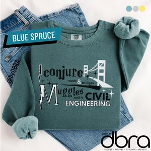 Könnte beinhalten: Ein blaugrünes Sweatshirt mit dem Text "I conjure roads & bridges, Muggles call it CIVIL ENGINEERING" und einer Brückengrafik. Das Sweatshirt ist auf Jeans und einem passenden Scrunchie gefaltet.