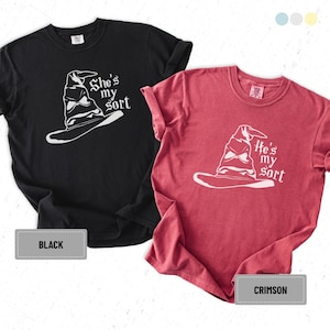 Könnte beinhalten: Zwei T-Shirts, eines in Schwarz und eines in Karmesinrot, jeweils mit einer weißen Grafik eines Sortierhuts. Das schwarze Shirt trägt die Aufschrift "She's my sort", während das rote Shirt "He's my sort" trägt.