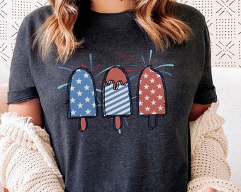 May include: A dark gray t-shirt with a graphic of three ice cream pops decorated with red, white, and blue stars and stripes. The ice cream pops are surrounded by red and blue fireworks.
