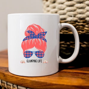 May include: White ceramic mug with a graphic of a woman with a red bun, blue and red plaid headband, and blue and red plaid sunglasses. The text "GLAMPING LIFE" is below the graphic, with two small camper icons on either side.