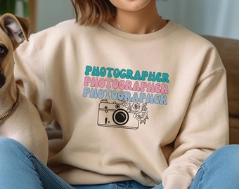 May include: A beige sweatshirt with the text "PHOTOGRAPHER" repeated three times in different colors. A vintage camera with flowers is below the text.