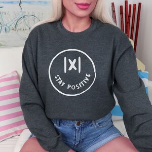 May include: Dark gray sweatshirt with a white circular graphic. The graphic features a stylized face with the text "STAY POSITIVE" in a circular pattern. The sweatshirt is a crew neck style.
