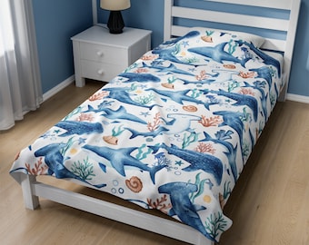 May include: A white twin-sized bedspread with a blue and white watercolor pattern of sharks, seashells, and coral. The bedspread is on a white bed frame with a blue headboard.