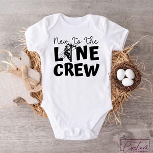 May include: White baby bodysuit with the text "New To The Line Crew" in black. The bodysuit features a graphic of a lineman on a power line. The bodysuit is displayed on a wooden surface with a decorative bunny and a nest with eggs.