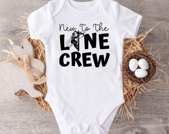 May include: White baby bodysuit with the text "New To The Line Crew" in black. The bodysuit features a graphic of a lineman on a power line. The bodysuit is displayed on a wooden surface with a decorative bunny and a nest with eggs.