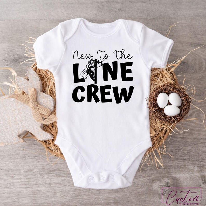 May include: White baby bodysuit with the text "New To The Line Crew" in black. The bodysuit features a graphic of a lineman on a power line. The bodysuit is displayed on a wooden surface with a decorative bunny and a nest with eggs.