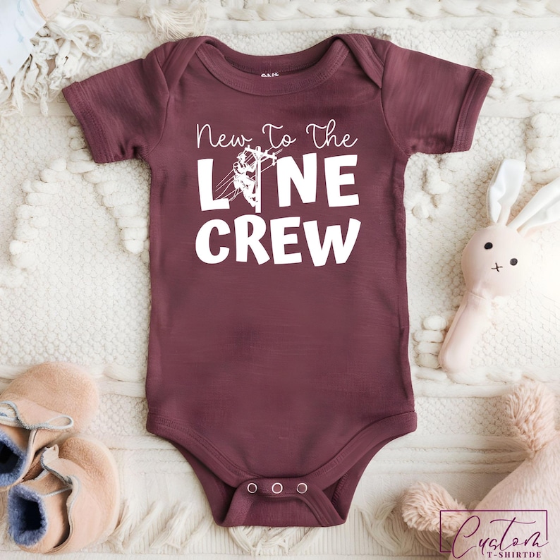 May include: Burgundy baby onesie with the text "New To The Line Crew" in white. The design includes an illustration of a lineman on a power pole. The onesie is surrounded by a soft, white blanket and baby accessories.