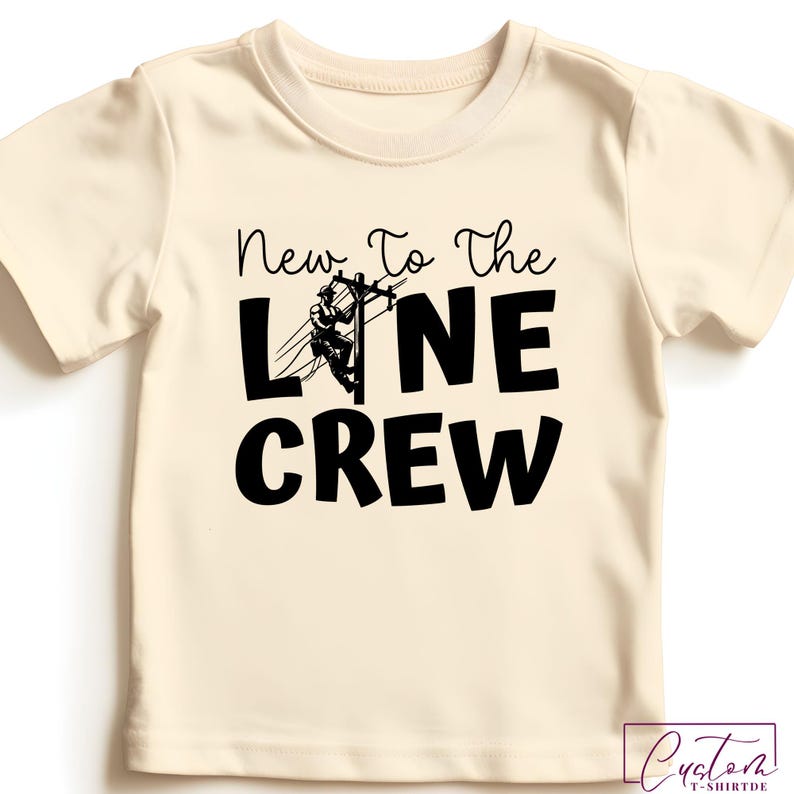 May include: Cream-colored t-shirt with the text "New To The LINE CREW" in black. The design includes a graphic of a lineman on a power line. The shirt is a short-sleeved crew neck.