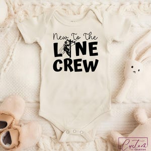 May include: Cream-colored baby onesie with the text "New To The Line Crew" in black. A graphic of a lineman working on a power line is incorporated into the text. The onesie has short sleeves and snap closures.