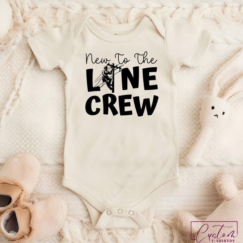 May include: Cream-colored baby onesie with the text "New To The Line Crew" in black. A graphic of a lineman working on a power line is incorporated into the text. The onesie has short sleeves and snap closures.