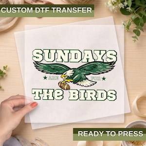 May include: A ready-to-press DTF transfer featuring the text "SUNDAYS ARE FOR THE BIRDS" in green and white, with an eagle graphic holding a football. The transfer is on a transparent sheet, ideal for apparel customization.