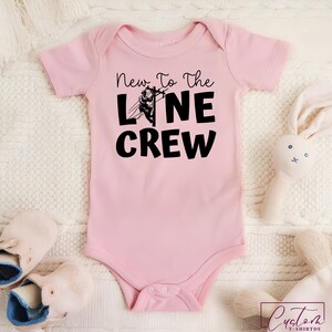 May include: A light pink baby onesie with the text "New To The Line Crew" in black. The design includes a graphic of a lineman working on a power line. The onesie is surrounded by a white blanket, a pair of baby shoes, and a stuffed bunny.
