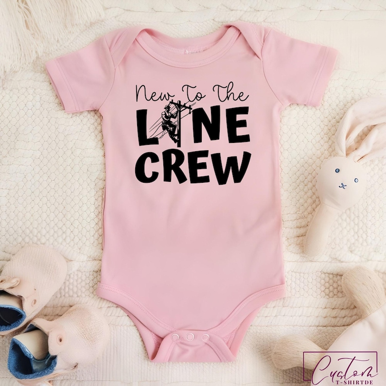 May include: A light pink baby onesie with the text "New To The Line Crew" in black. The design includes a graphic of a lineman working on a power line. The onesie is surrounded by a white blanket, a pair of baby shoes, and a stuffed bunny.