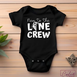 May include: Black baby bodysuit with the text "New To The Line Crew" in white. The design includes a graphic of a lineman on a power line. The bodysuit is made of soft material and has snap closures.