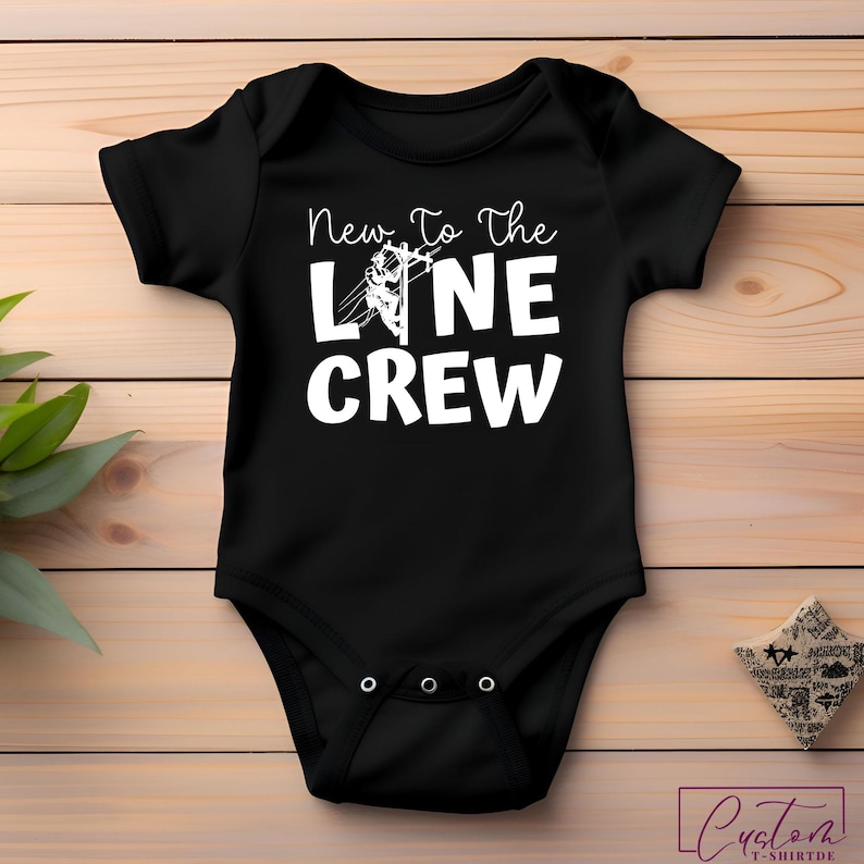 May include: Black baby bodysuit with the text "New To The Line Crew" in white. The design includes a graphic of a lineman on a power line. The bodysuit is made of soft material and has snap closures.
