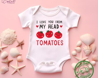 May include: White baby bodysuit with the text "I LOVE YOU FROM MY HEAD TOMATOES" in black and red. The design includes three smiling tomato illustrations. The bodysuit has short sleeves and snap closures.