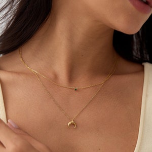 Puede incluir: Un collar de cadena de oro con un colgante de media luna y una pequeña piedra preciosa verde. El collar está superpuesto con una cadena delicada con una sola piedra preciosa verde.
