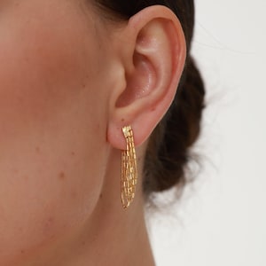 May include: Gold dangle earrings with a layered chain design. The earrings are shown in a close-up view on a person's ear.