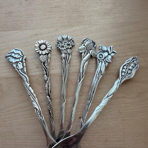 May include: A collection of antique silver spoons with ornate floral designs on the handles. Each spoon features a different flower, including a daisy, carnation, and lily of the valley. The spoons are arranged on a light brown wooden surface.