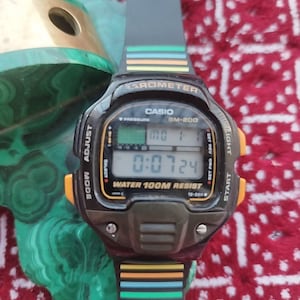 Może przedstawiać: Czarny zegarek cyfrowy Casio BM-200 z czarnym paskiem i kolorowymi paskami. Tarcza zegarka wyświetla godzinę, datę i inne funkcje. Posiada barometr i wodoszczelność do 100 metrów.