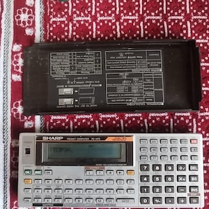 Puede incluir: Una calculadora de bolsillo Sharp PC-1475 plateada y gris con una cubierta protectora negra. La calculadora tiene numerosos botones y una pantalla que muestra texto. La cubierta tiene instrucciones impresas.