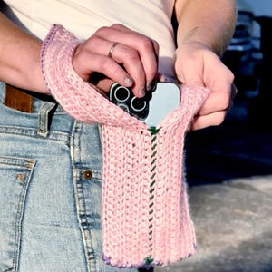 May include: A light pink knitted phone case with a strap. The case is made of a chunky knit with a green and purple trim. The phone case is holding a phone with multiple camera lenses.