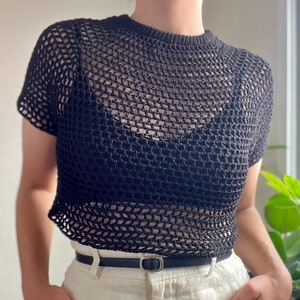 Puede incluir: Un top de crochet negro de corte holgado y mangas cortas. El top tiene un escote cuadrado y un delicado patrón de tejido abierto. Un cinturón negro con hebilla plateada se lleva sobre el top.