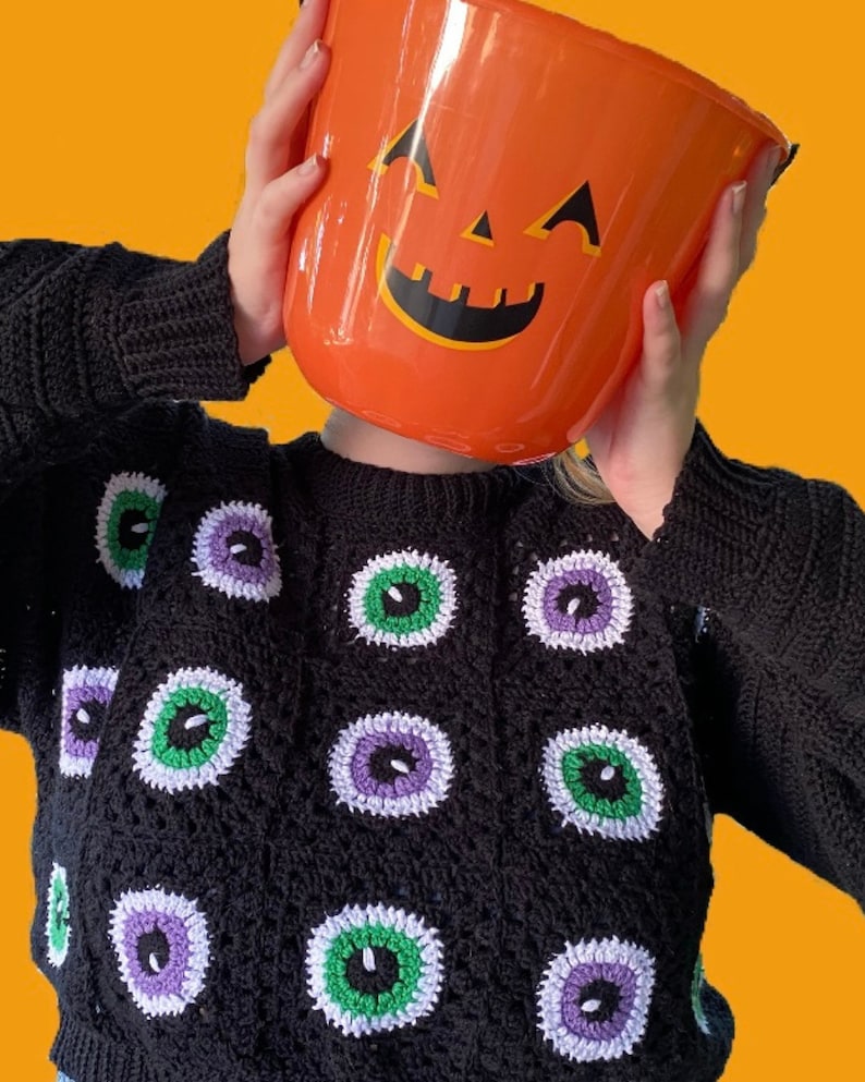 Puede incluir: Un su&eacute;ter de crochet negro con un patr&oacute;n de ojos coloridos. El su&eacute;ter lo lleva una persona que sostiene un cubo de calabaza naranja brillante frente a su cara.