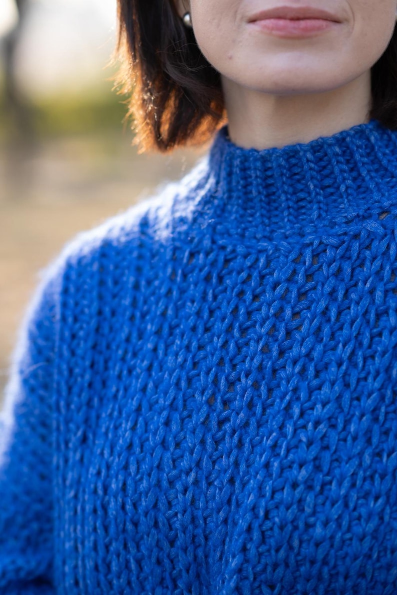 May include: A close-up of a blue knitted sweater with a high neck. The sweater is made of a soft, textured yarn.