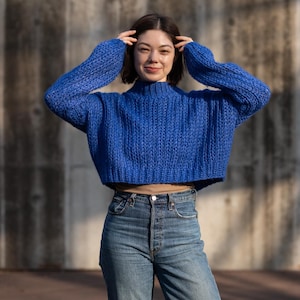 May include: A woman wearing a blue cropped sweater and blue jeans. She is standing in front of a concrete wall.