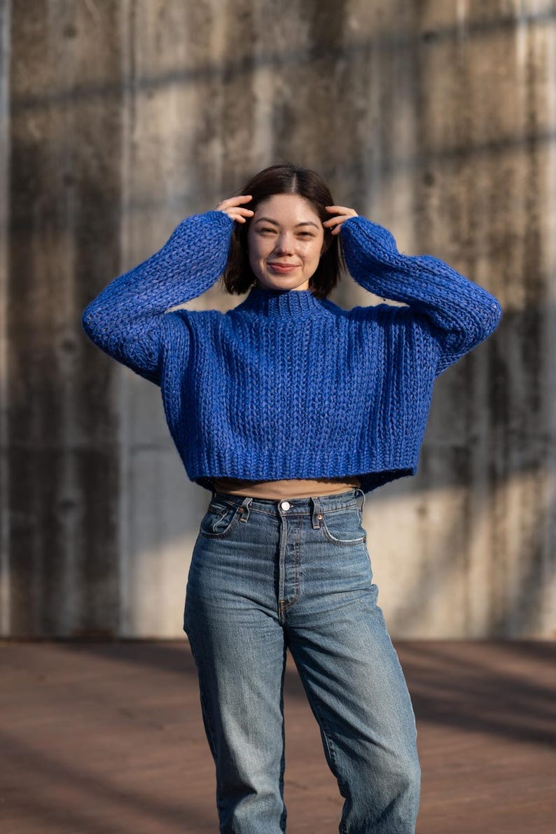 May include: A woman wearing a blue cropped sweater and blue jeans. She is standing in front of a concrete wall.