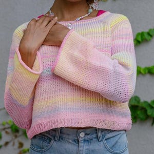 Può includere: Un maglione dai colori pastello con scollo tondo e maniche lunghe a campana. La maglia presenta una sfumatura di rosa, giallo e blu. Il maglione è abbinato a jeans di denim chiaro.