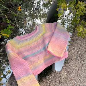 Peut inclure: Un pull aux couleurs pastel avec un col rond et des manches longues. Le pull présente un dégradé de teintes roses, jaunes et bleues. Les manches ont une conception évasée plus large. Le pull est en matière tricotée douce.