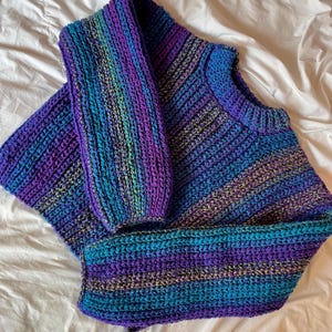 May include: A handmade, multi-colored sweater in shades of purple, blue, and teal. The sweater features a ribbed knit design and long sleeves. The garment is displayed on a white surface.