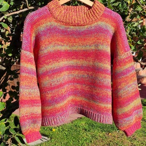 May include: A colourful striped knitted jumper with a high neck and long sleeves. The jumper is made of a soft, chunky yarn and features a variety of shades of pink, orange, yellow, and purple.
