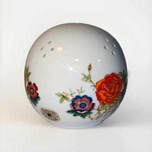 May include: A spherical white porcelain shaker featuring a colorful floral pattern with orange, red, and blue flowers and green leaves. The top surface has small perforated holes for dispensing, and a circular sticker is visible on the side.