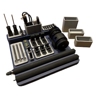 Puede incluir: Un organizador azul con varios compartimentos para guardar pequeñas herramientas y accesorios. Incluye un engranaje negro, un organizador de cables gris y varios compartimentos negros y grises. El organizador está sobre una superficie blanca.