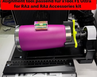 Könnte beinhalten: Ein lila-silbernes Ausrichtungswerkzeug für den xTool F1 Ultra Lasergravierer. Das Werkzeug befindet sich auf einer schwarzen Basis mit einem Lineal und einer Wasserwaage. Der Text auf der Basis lautet "Drehen zum Zurücksetzen", "Schlüssel prüfen" und "Schutz nach unten ziehen".