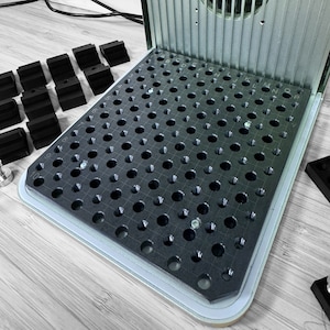 Op de afbeelding: Een zwart-wit raster met gaten om kleine voorwerpen vast te houden. Het raster is gelabeld met letters en cijfers. Het raster maakt deel uit van een grotere machine.