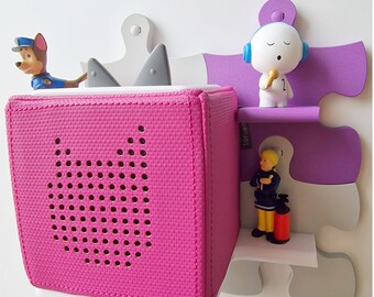 May include: A pink sound cube with a speaker grille and a white trim. The cube is attached to a white and purple puzzle-shaped wall system. The text "Beliebig erweiterbar: Steckbar ohne Schrauben" is visible on the wall system.