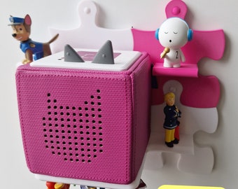 May include: A pink cube-shaped speaker with a speaker grille on the front. The speaker is attached to a white jigsaw piece wall mount. The wall mount has a pink shelf with a white figure holding a microphone. The text on the yellow sticker reads "Beliebig erweiterbar. Steckbar ohne Schrauben".