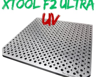 Puede incluir: Una placa de plantilla de metal plateado con numerosos agujeros espaciados uniformemente. El texto "THE JIGMAKER FOR XTOOL F2 ULTRA UV" se muestra en verde y rojo. The Jigmaker F2 está impreso en la esquina inferior derecha.